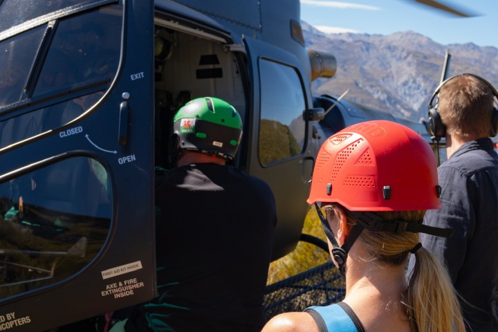 People boarding a helicopter on a grassy area with mountains in the background.