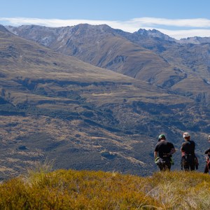Three hikers in gear overlook a vast mountainous landscape.