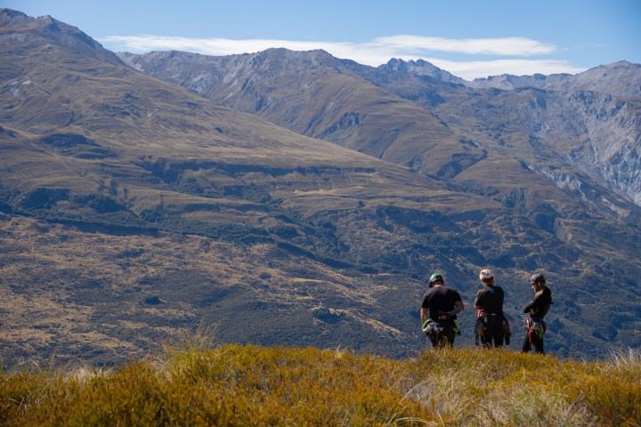 Three hikers in gear overlook a vast mountainous landscape.