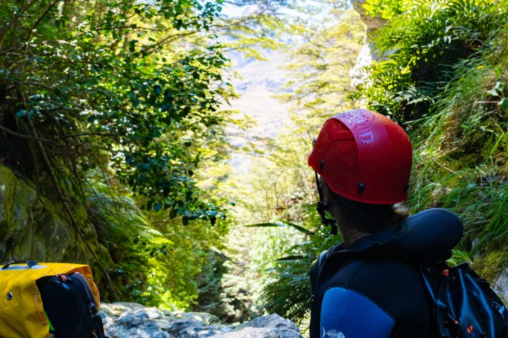 Person in helmet and wetsuit sitting among rocks and foliage, looking at a lush canyon.