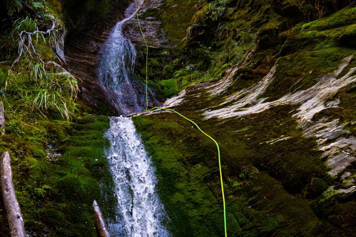 Person rappelling down a waterfall in a lush, narrow canyon.