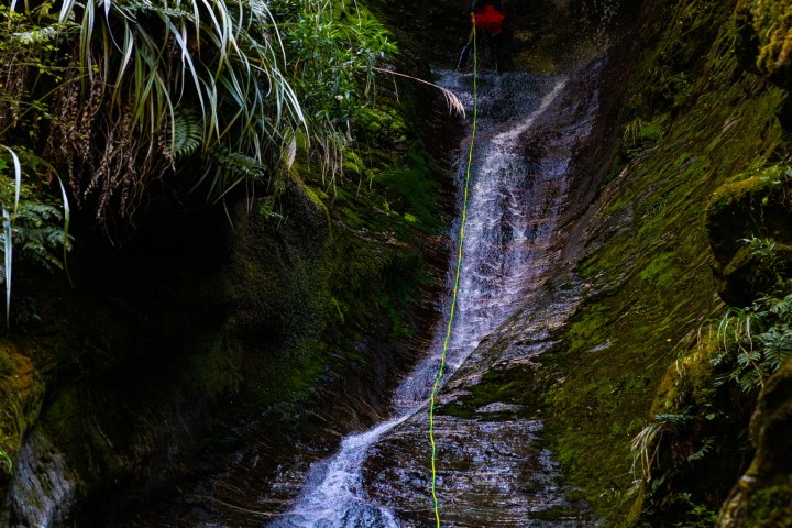 Person rappelling down a lush, narrow canyon with a waterfall.