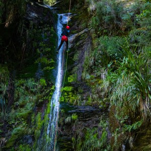 Person rappelling down a waterfall in a lush, rocky environment.