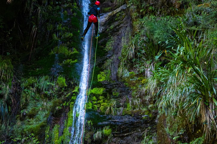 Person rappelling down a waterfall in a lush, rocky environment.
