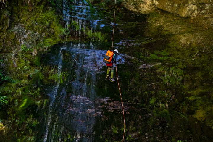 Two individuals rappel down a mossy cliff with a small waterfall cascading beside them.