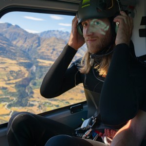 Person in helmet sitting in helicopter with mountain view outside.