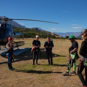 Group in outdoor gear stands near helicopter on grassy field.