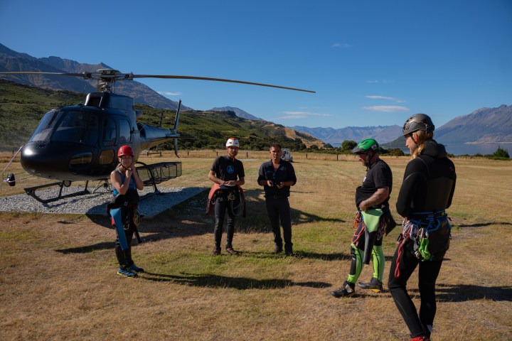 Group in outdoor gear stands near helicopter on grassy field.
