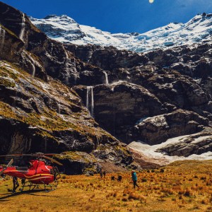 Red helicopter on grassy field near snowy mountains and waterfalls.