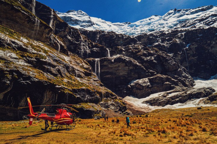Red helicopter on grassy field near snowy mountains and waterfalls.