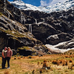 Two people stand in front of a rocky mountain with snow and waterfalls.