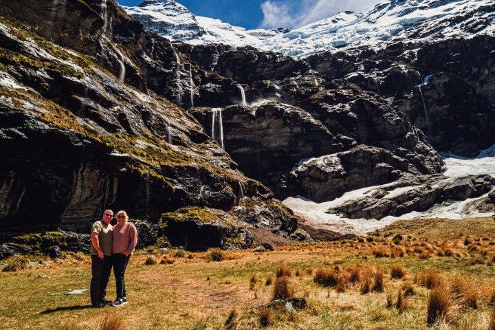 Two people stand in front of a rocky mountain with snow and waterfalls.