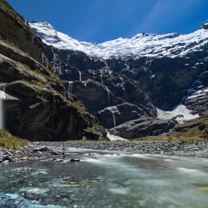 Waterfalls cascade into a mountain stream with snow-capped peaks in the background under a clear blue sky.