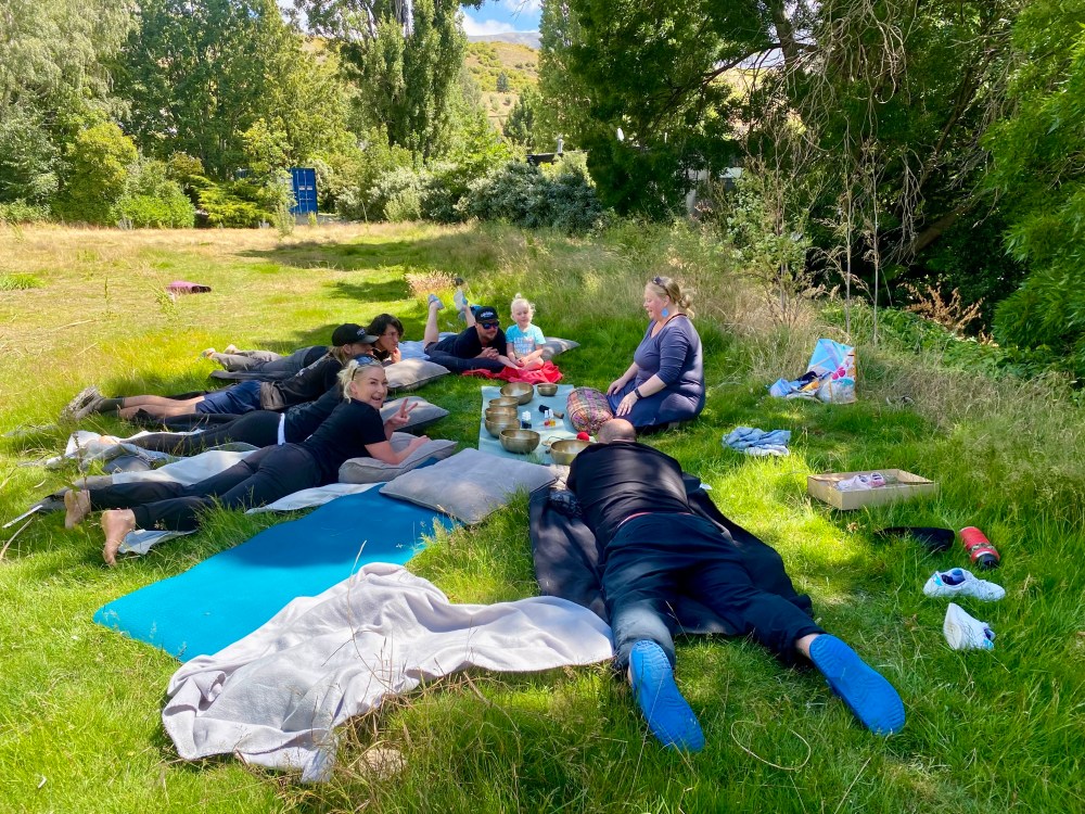 Group of people relaxing on mats in a grassy field under trees.