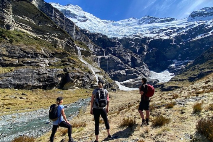 Three hikers view a snow-capped mountain with waterfalls.