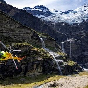 Yellow helicopter flying near waterfalls in a mountainous landscape with snow-capped peaks.