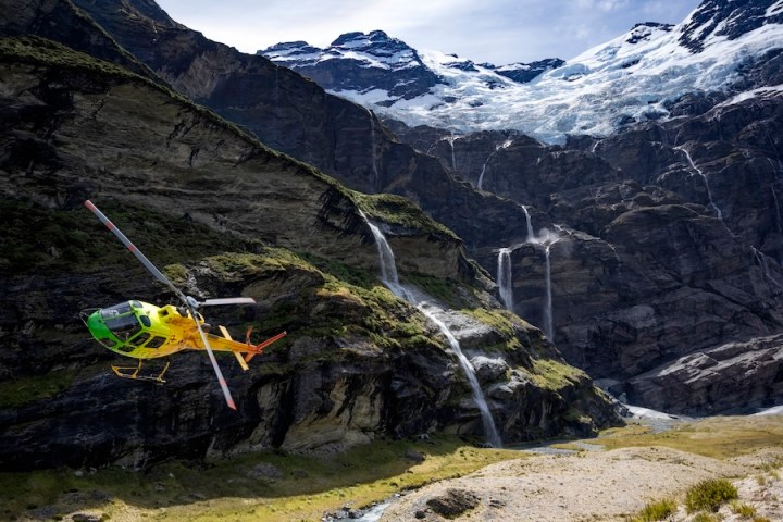Yellow helicopter flying near waterfalls in a mountainous landscape with snow-capped peaks.