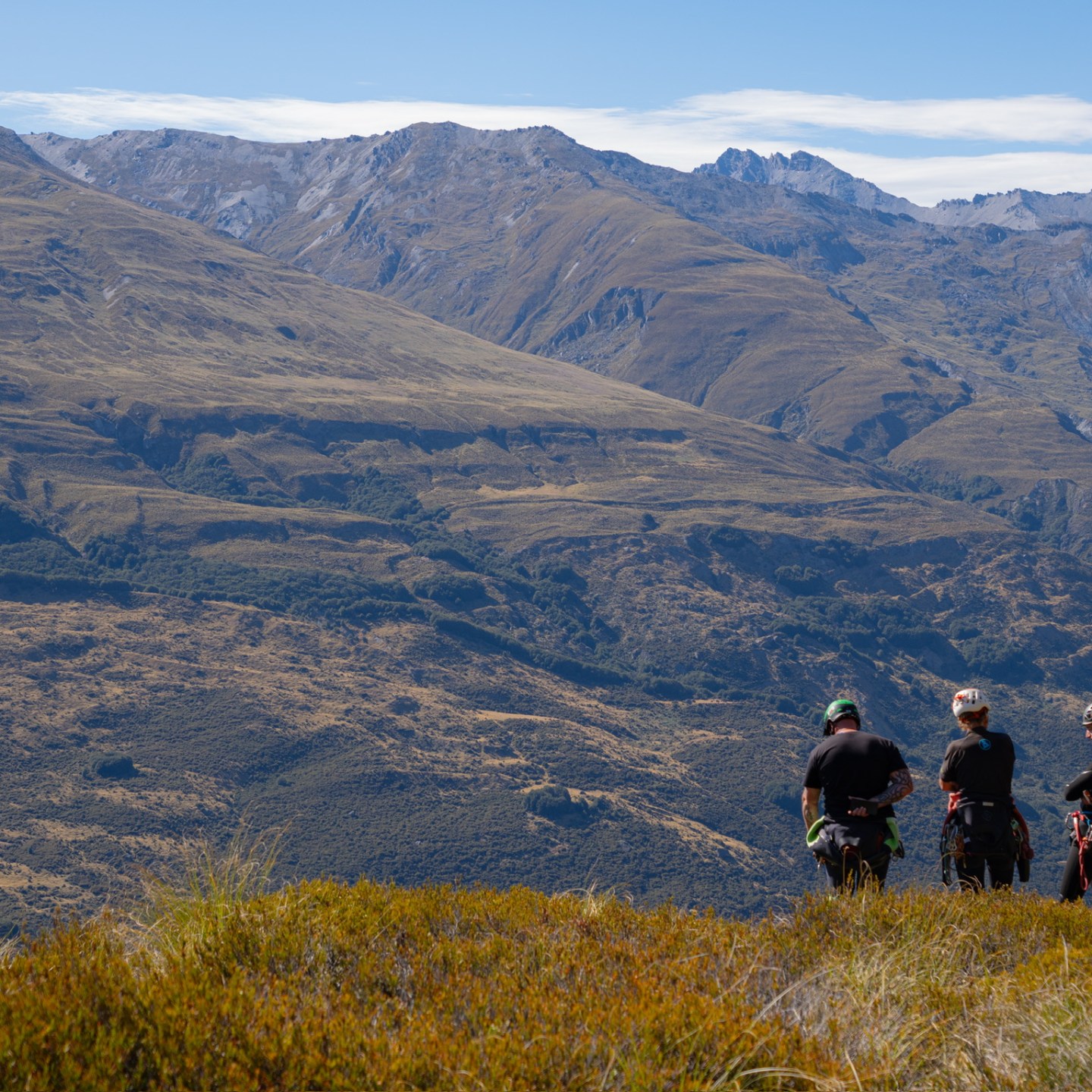 Three people stand on a grassy hill with mountains in the background.