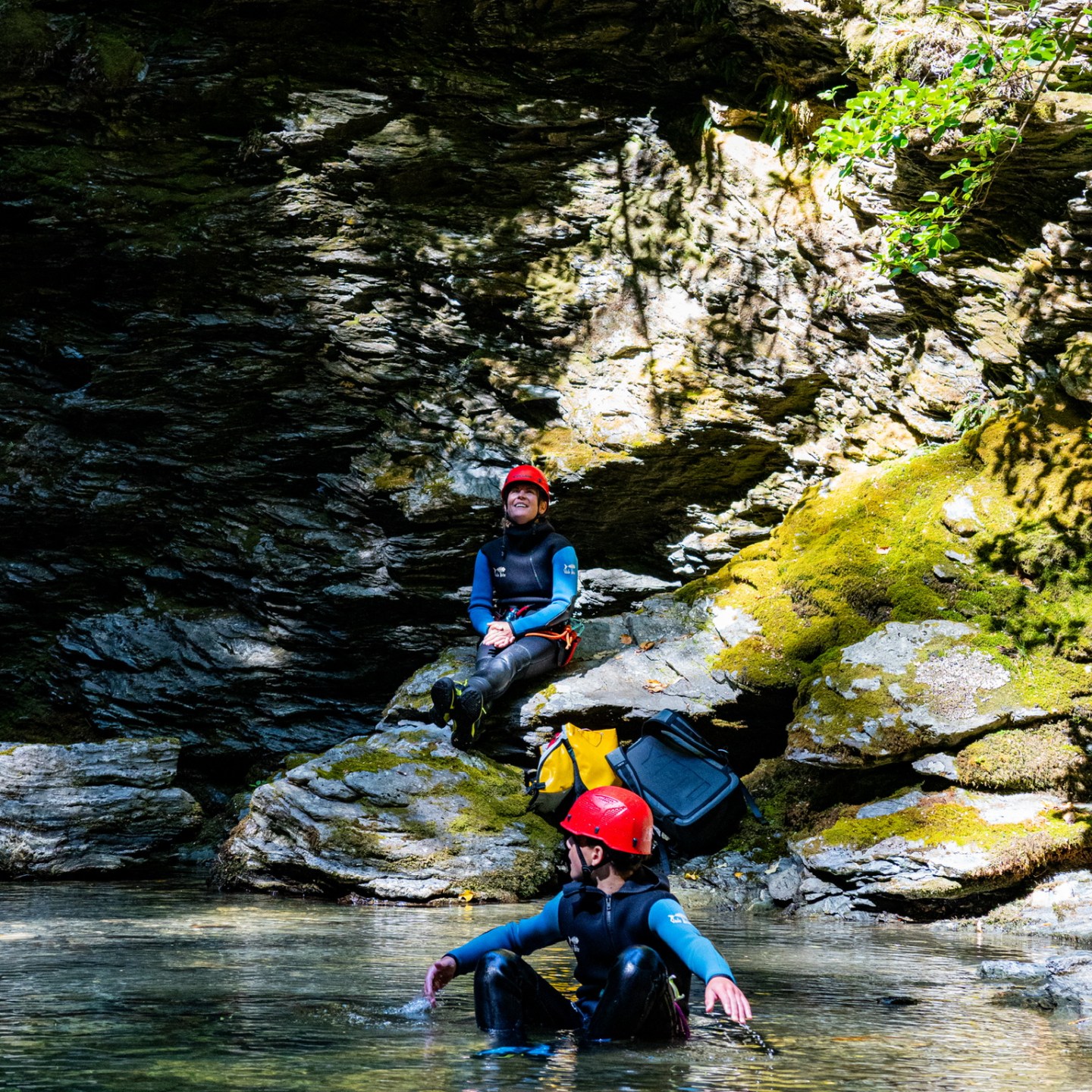 Two people in wetsuits and helmets by rocky water's edge.