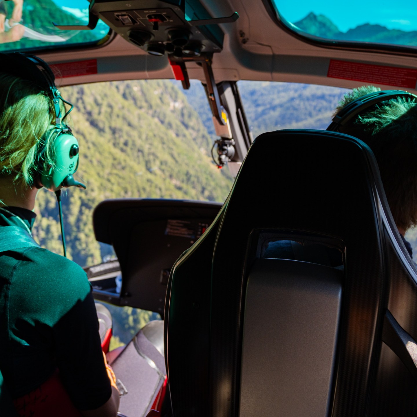 Three people inside a helicopter flying over mountainous terrain.