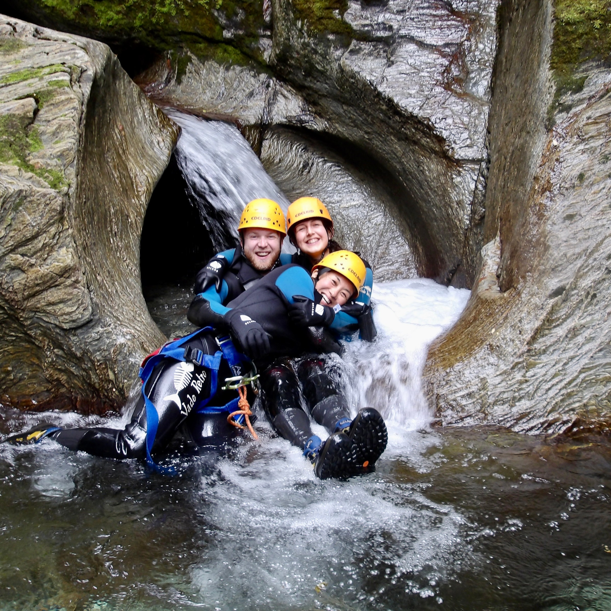 Three people in helmets smiling while seated in a rocky water slide with flowing water.