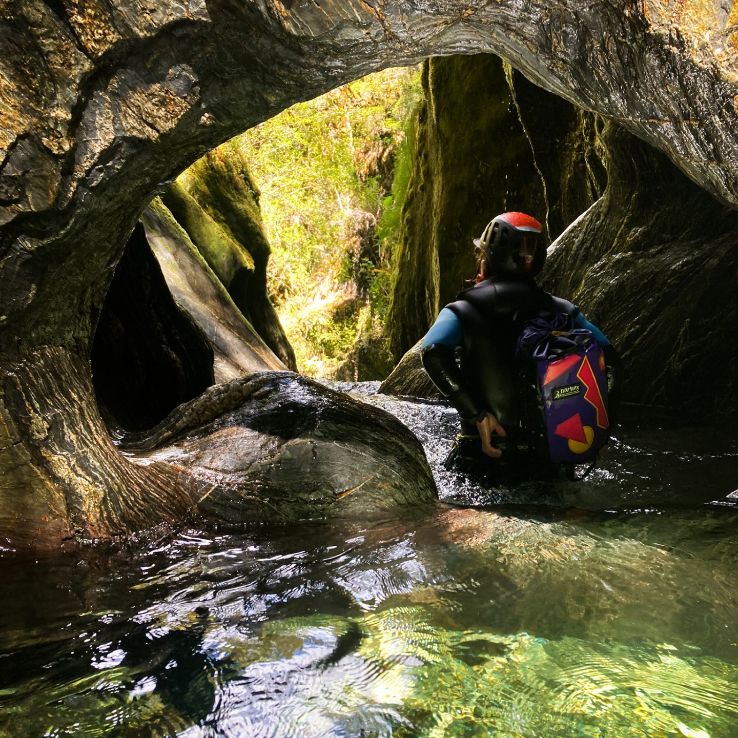 Person in wetsuit exploring rocky cave with clear water and sunlight.