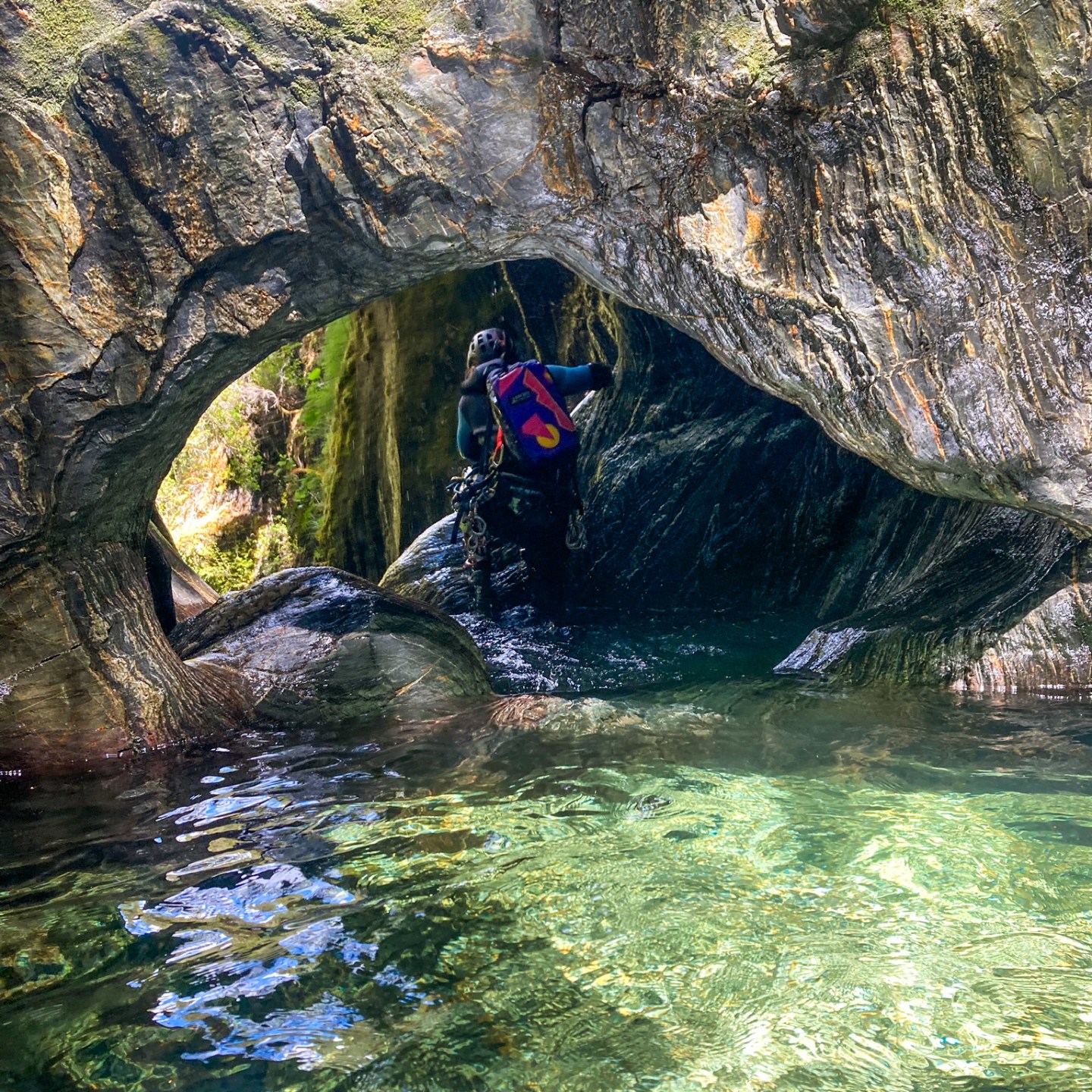 Person with backpack exploring cave with clear water and mossy rock walls.