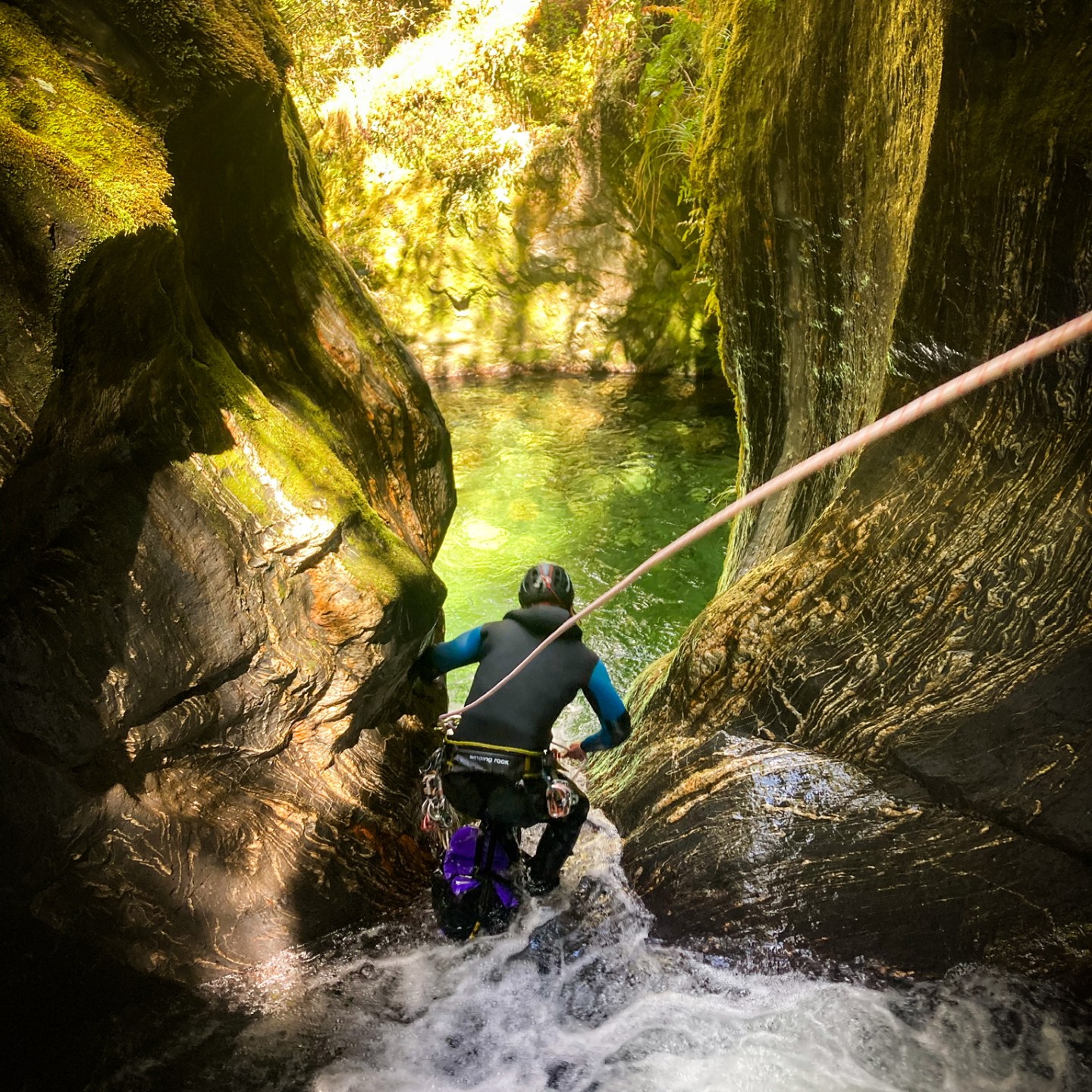 Person canyoning down a waterfall into a clear pool in a rocky gorge.