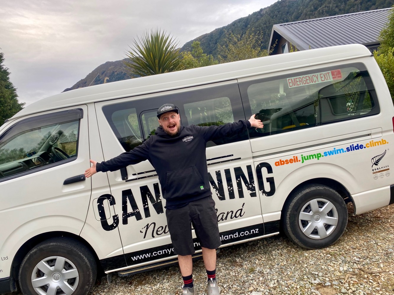 Person posing with arms outstretched in front of a van labeled 'Canyoning New Zealand' in a scenic area.
