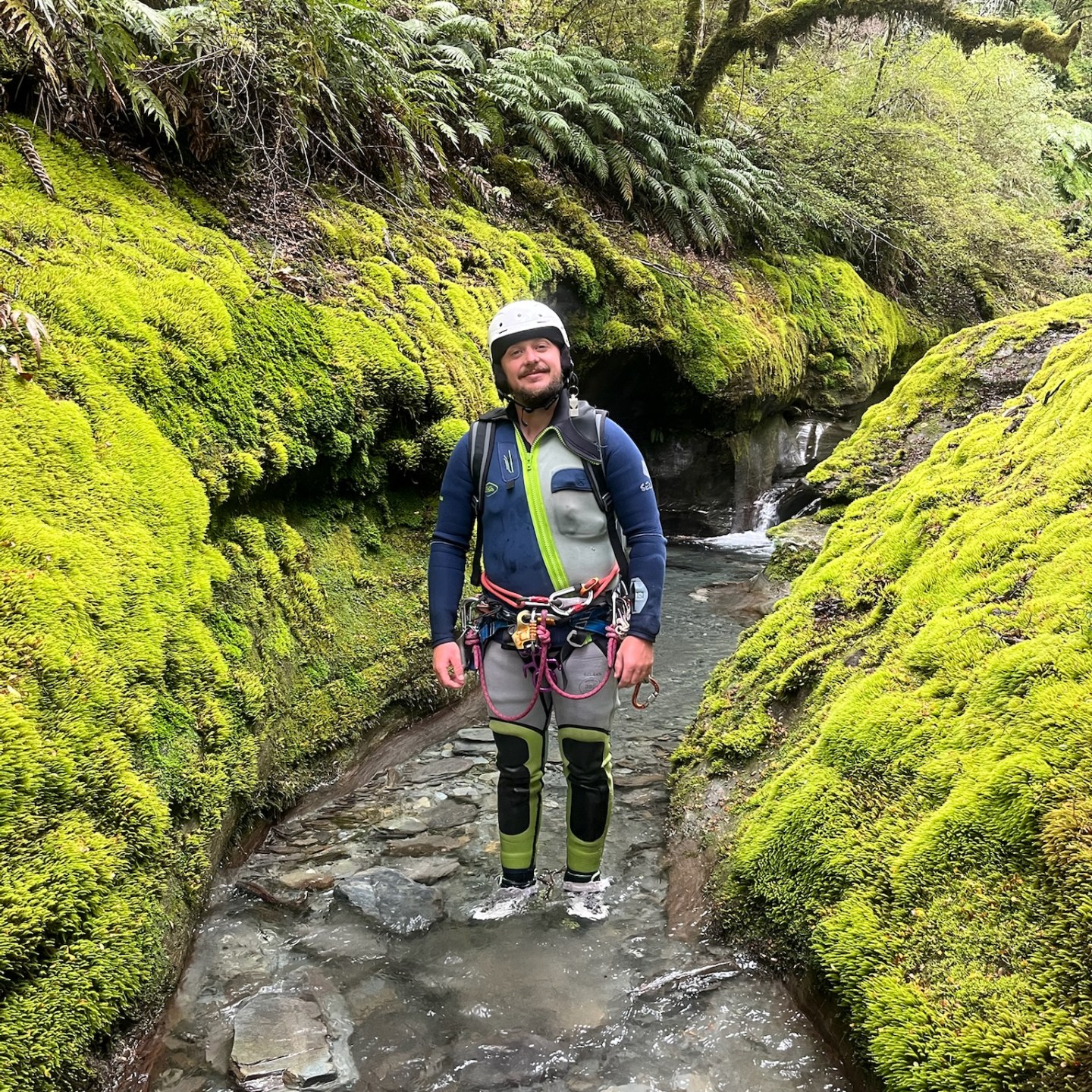 Person in wetsuit and helmet stands in mossy canyon creek.