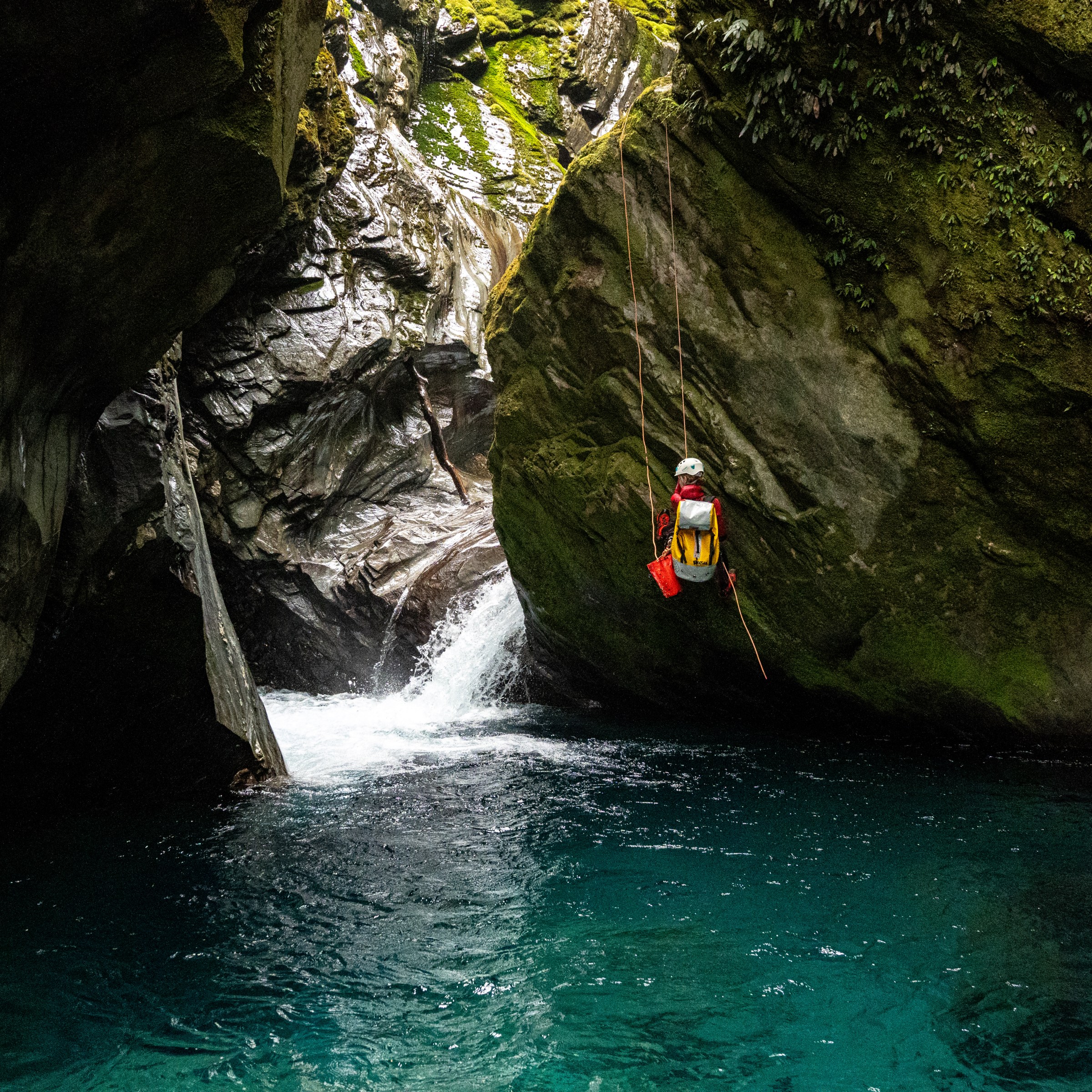Canyoneer rappelling down a mossy rock into a blue pool.