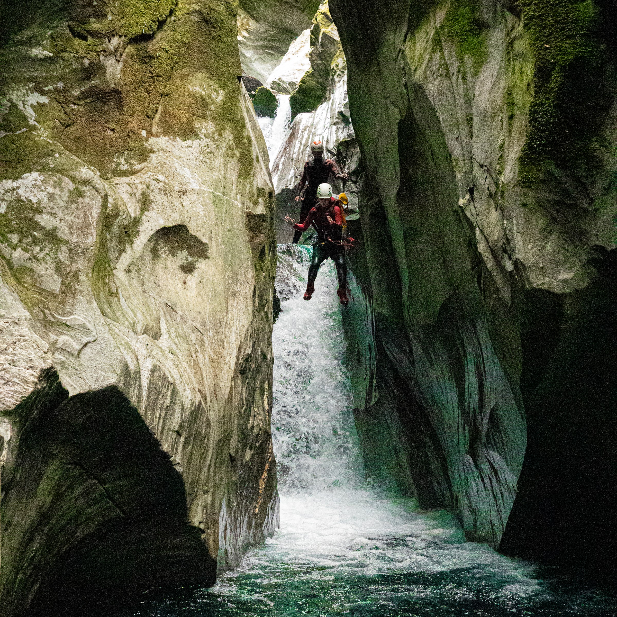 Two people canyoning down a narrow waterfall surrounded by tall rock walls.