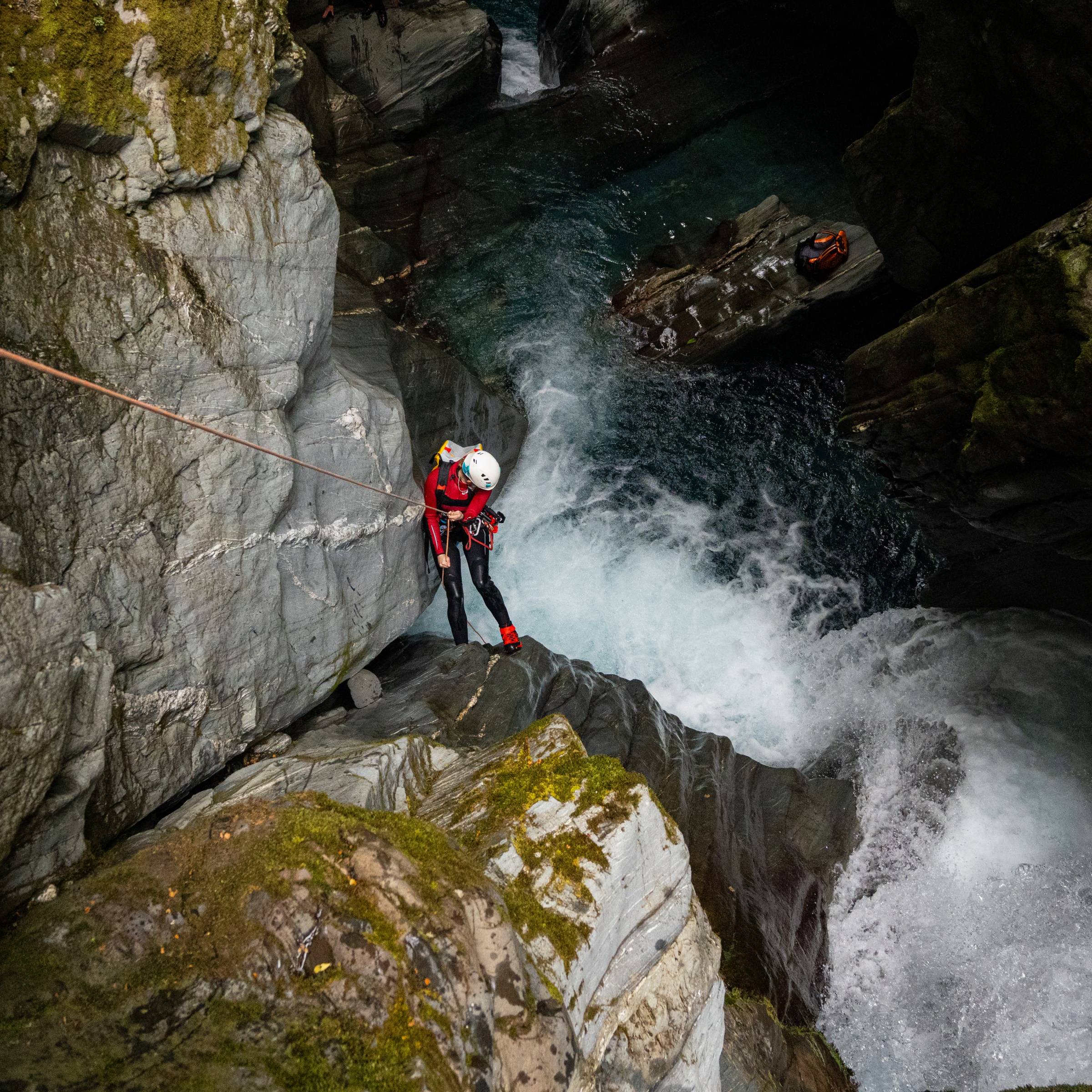 Person in red gear canyoning down a waterfall with a rope in a rocky gorge.
