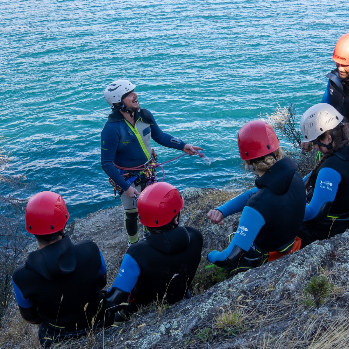 Group wearing helmets and wetsuits sitting on a cliff by water, listening to an instructor.