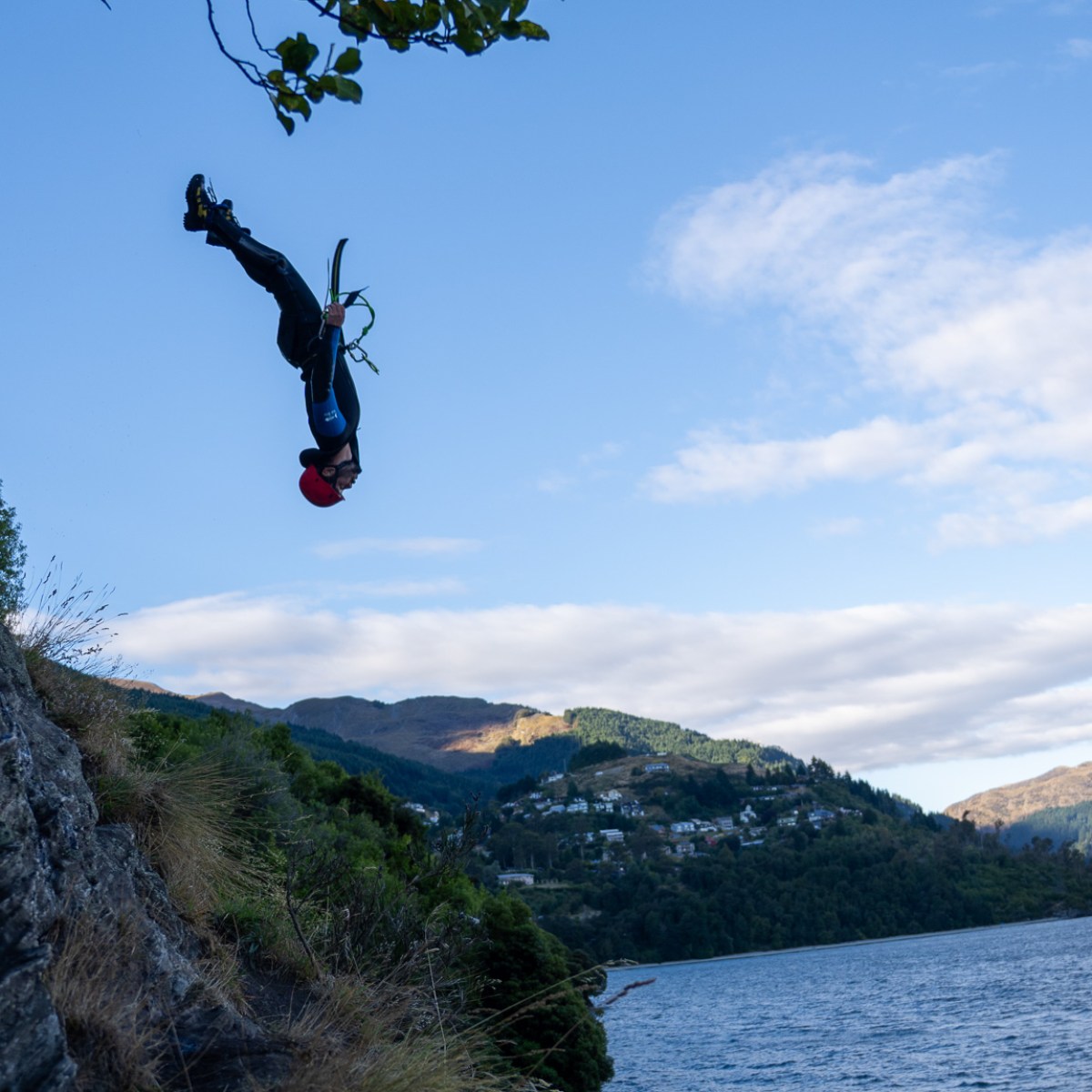 Person diving into lake from a cliff, surrounded by trees and mountains, under a blue sky.