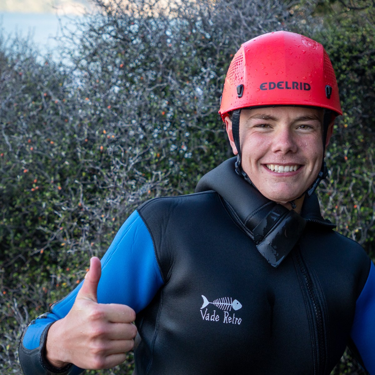Person in wetsuit and red helmet giving thumbs up outdoors.