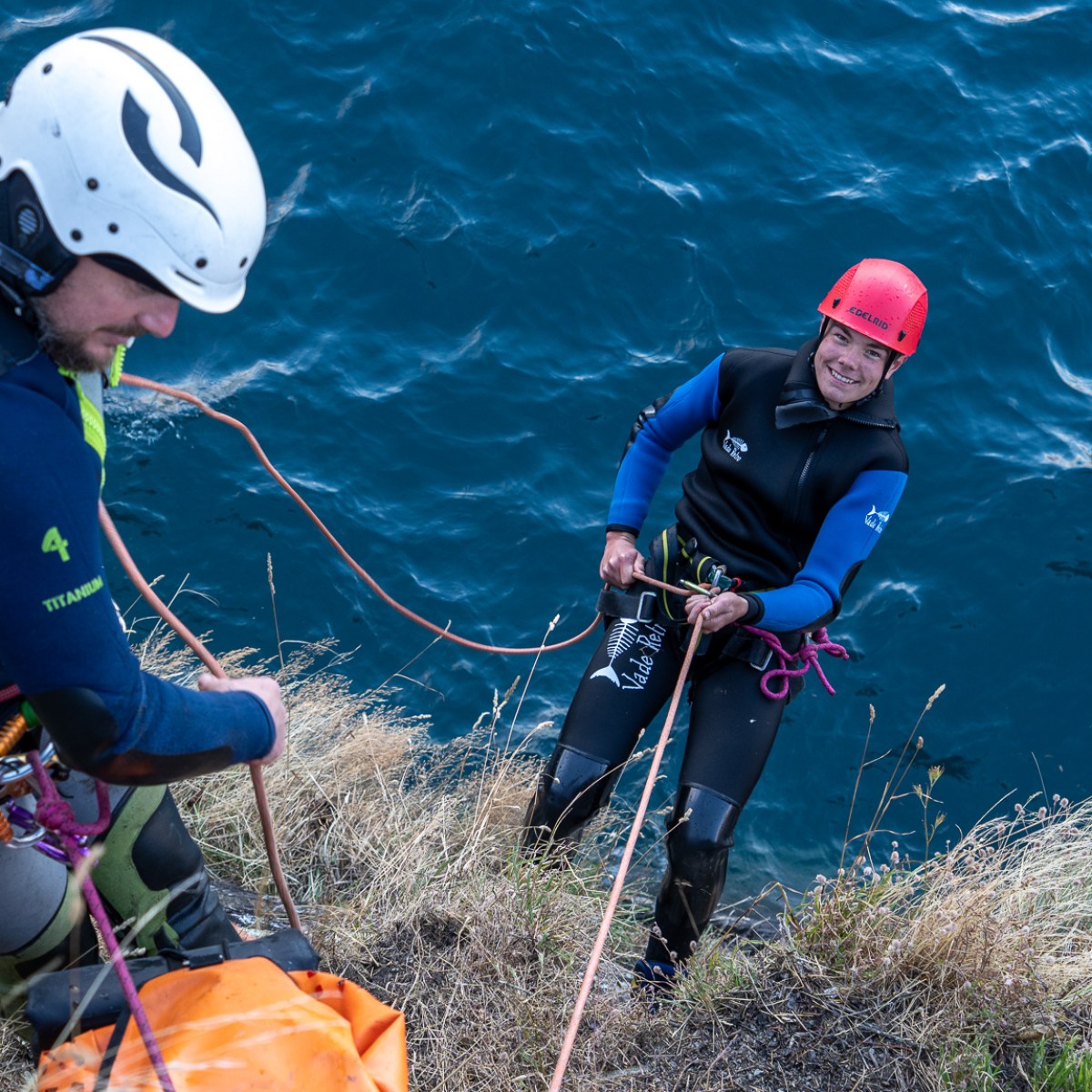Two people in helmets abseiling down a grassy cliff by the sea.