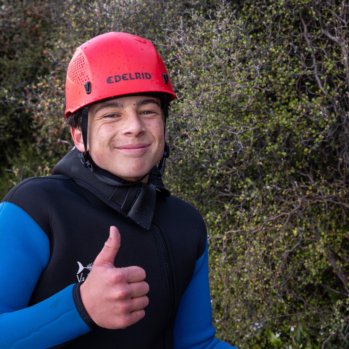Person in helmet and wetsuit giving a thumbs-up outdoors.
